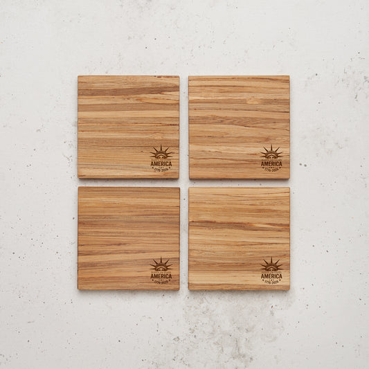 A set of four square coasters made of light brown, grained wood or bamboo arranged in a two-by-two grid on a textured grey surface. Each coaster features a small laser-engraved circular logo in the bottom right corner displaying the Statue of Liberty's crown, stars, and the text "AMERICA EST. 1776-2026".