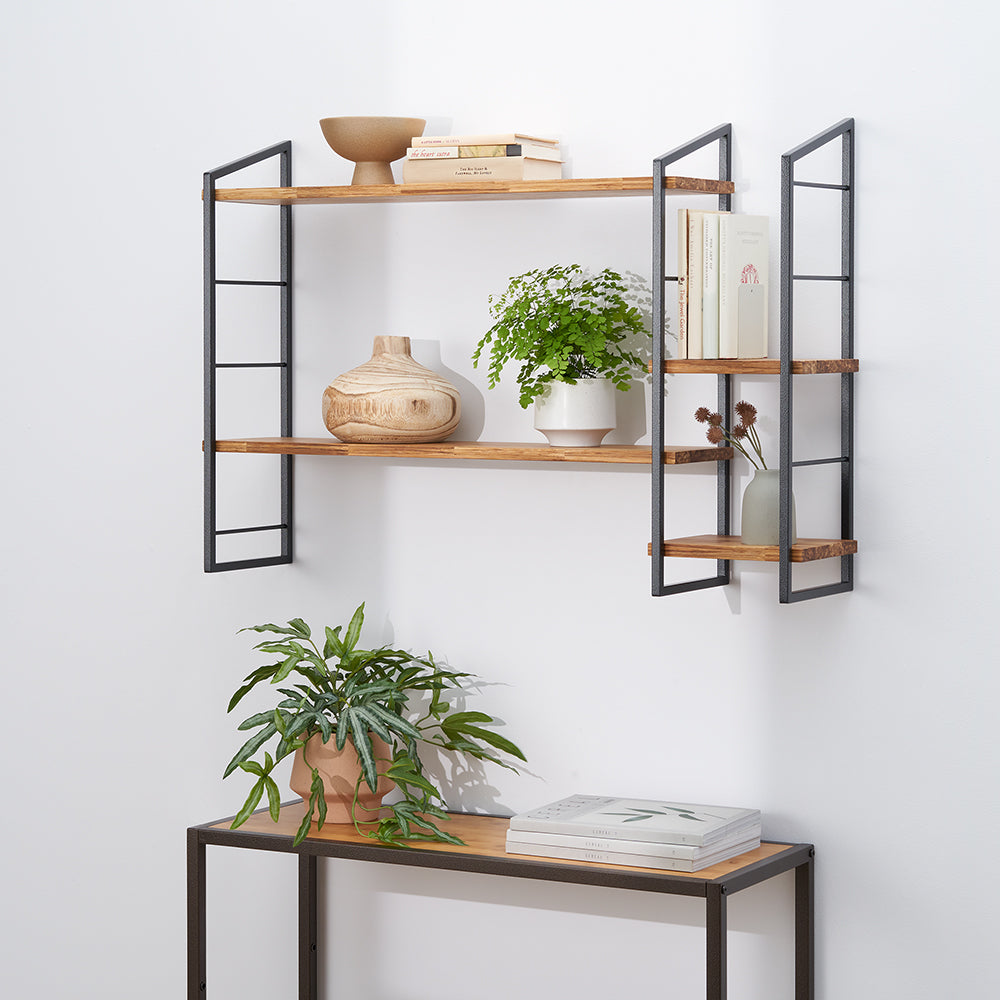 A pair of industrial-style, wall-mounted shelves featuring wood planks supported by black metal frames. The shelves are decorated with books, various potted plants, and wooden and ceramic vases, with a matching console table and plant below.
