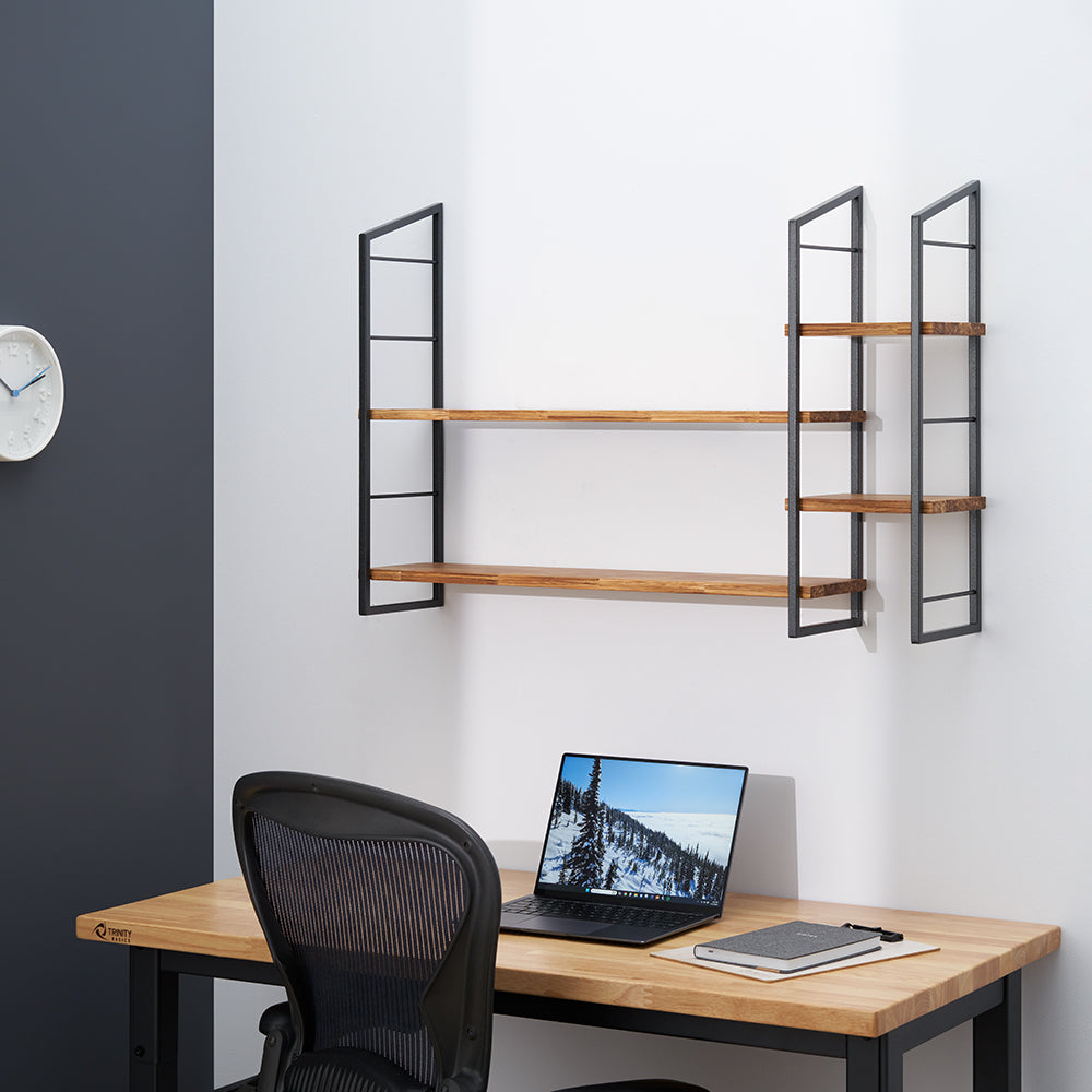 A modern home office setup featuring a wooden desk, an ergonomic office chair, and a laptop. Above the desk is a wall-mounted shelving unit with three natural wood shelves supported by black metal frames.