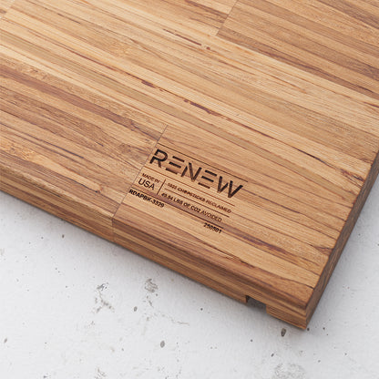A close-up shot of a light brown, rectangular cutting board made of many small, vertically aligned wooden blocks, likely bamboo. Engraved into the wood are the words "RENEW," "MADE IN USA," "1022 CHOPSTICKS RECLAIMED," and "49.94 LBS OF CO2 AVOIDED," along with some serial numbers.