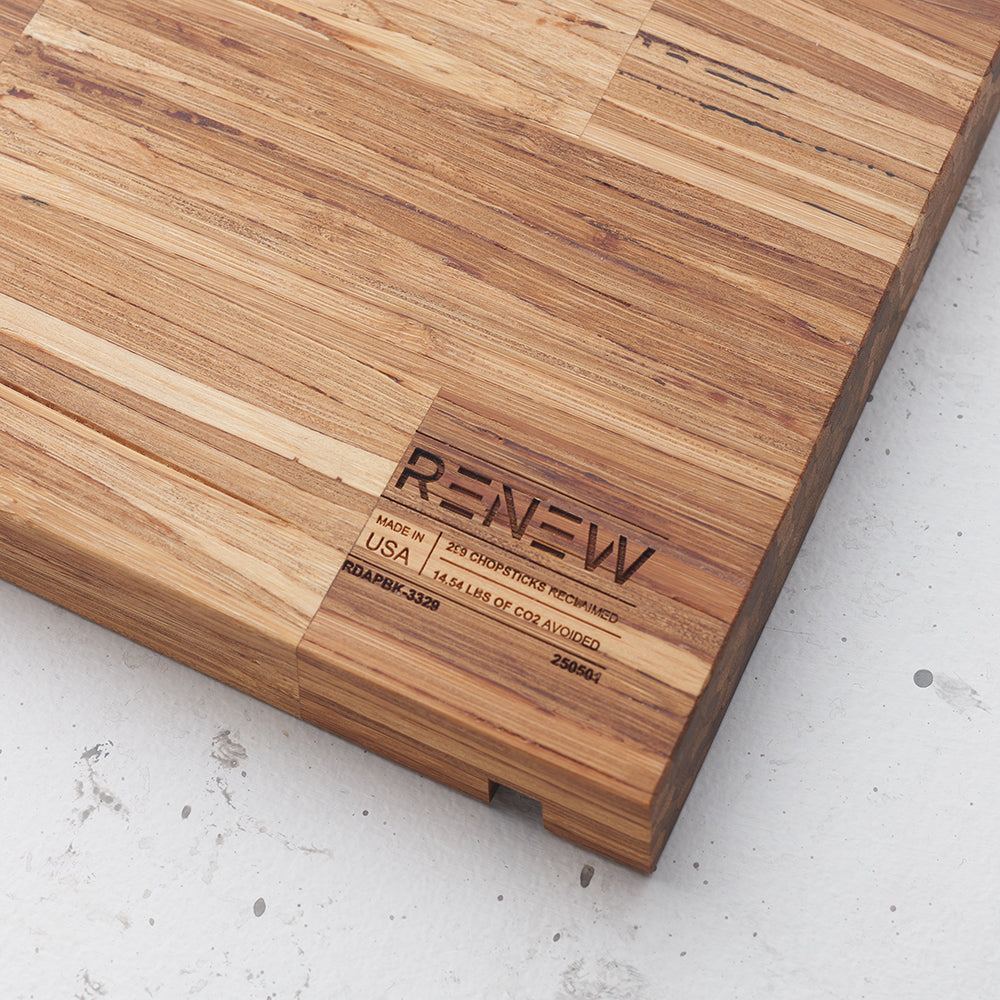 A close-up shot of a wooden cutting board, likely made of end-grain bamboo or reclaimed wood, shows a visible patchwork pattern of wood pieces. Engraved into the lower right corner are the words "RENEW," "MADE IN USA," "219 CHOPSTICKS RECLAIMED," and "14.54 LBS OF CO2 AVOIDED," along with other product codes.