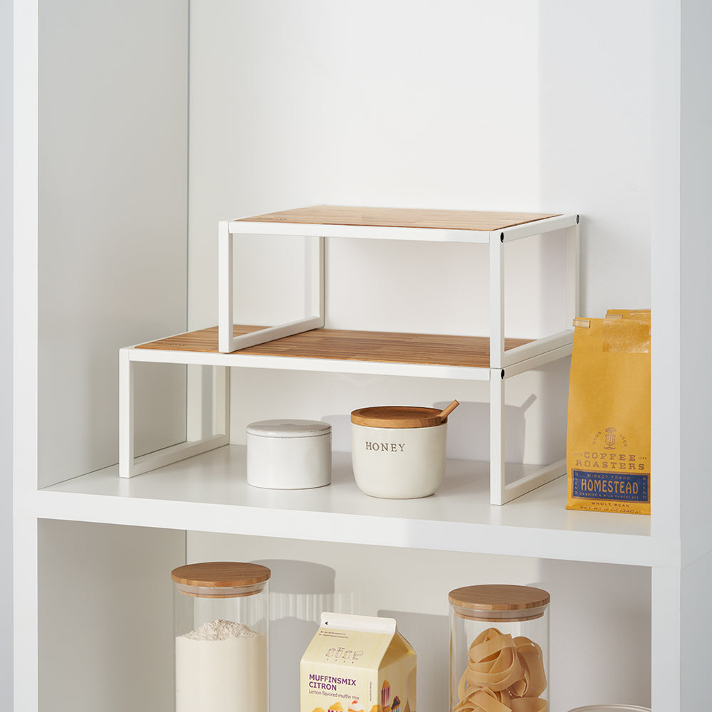 Two white metal shelf risers with bamboo tops are stacked in a tiered arrangement inside a white pantry or cabinet. They are used to organize kitchen items, including a ceramic honey jar, small containers, and a bag of coffee.