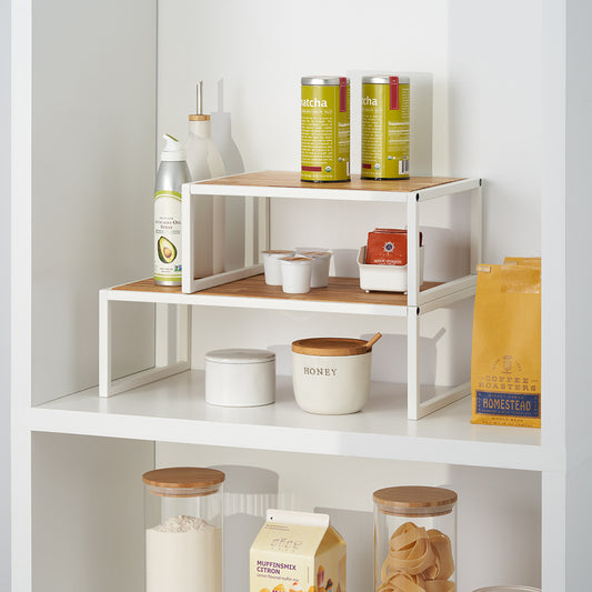 A white shelf features a two-tiered wooden and metal organizer holding matcha tins, coffee pods, and various pantry staples. The surrounding space is neatly arranged with a honey jar, avocado oil spray, and glass canisters containing dry goods.