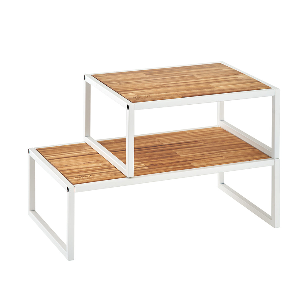 A two-tiered kitchen shelf features a white metal frame and natural wood surfaces with a butcher block pattern. The top shelf is smaller and nested over a larger bottom shelf to create an adjustable, tiered storage solution.