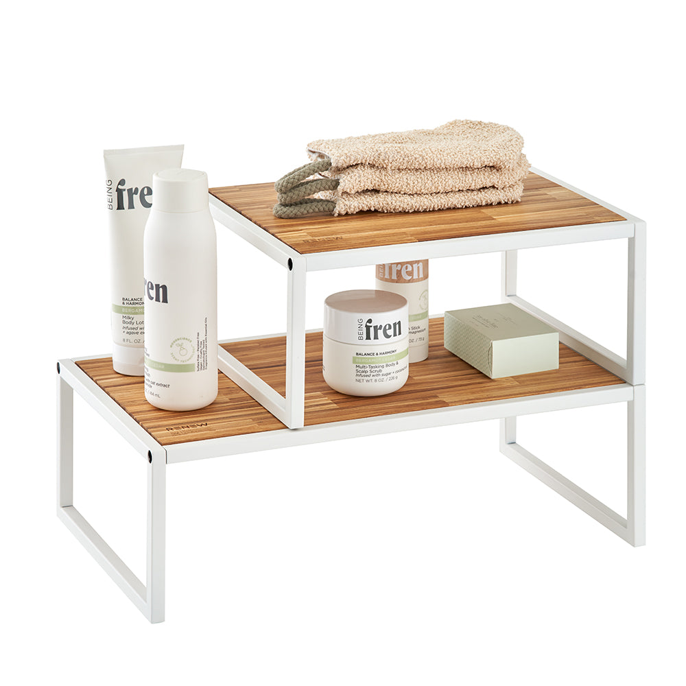 A two-tiered, white metal and wood organizing shelf holding various "Being Frenshe" brand skincare products and a stacked beige loofah. The modern, minimalist shelves are offset to create a staggered display, set against a clean white background.