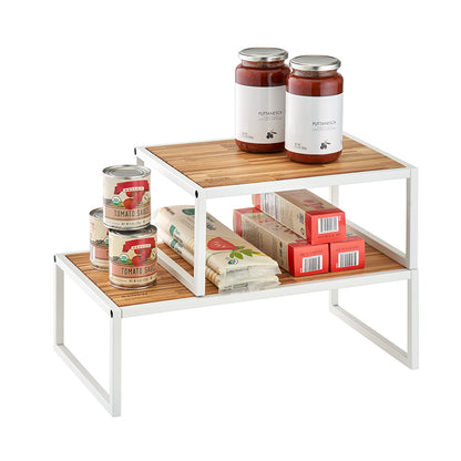 Two white metal pantry shelves with light wood surfaces are stacked in a tiered, offset arrangement to maximize storage space. The shelves are stocked with jars of puttanesca sauce, cans of tomato sauce, and boxes of pasta against a plain white background.
