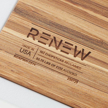 A close-up shot of a light-colored wooden surface engraved with the word "RENEW" in a bold, stylized font. The engraving includes details stating the product is "MADE IN USA" and was created using "502 CHOPSTICKS RECLAIMED," avoiding 53.75 lbs of CO2.