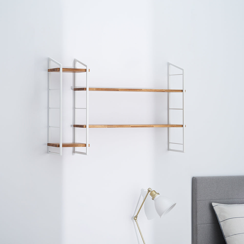A modern wall-mounted shelving unit features three wooden shelves supported by white metal ladder-style frames on both sides. Below the shelves, a brass and white desk lamp is visible in front of a gray upholstered headboard.