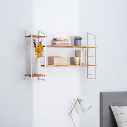 A modern, two-tiered wall shelf features light wood shelves and white wire metal frames mounted on a plain white wall above a bedroom setting. The shelves are styled with books, a small potted plant, a box, a candle, and a white vase holding yellow flowers.