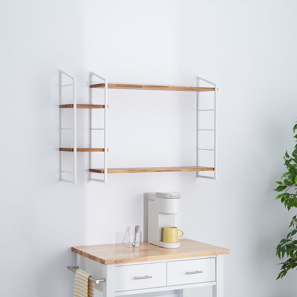 A set of white metal and natural wood wall-mounted shelves is installed above a white wooden kitchen cart with a butcher block top. A white single-serve coffee maker with a yellow mug is sitting on the cart.