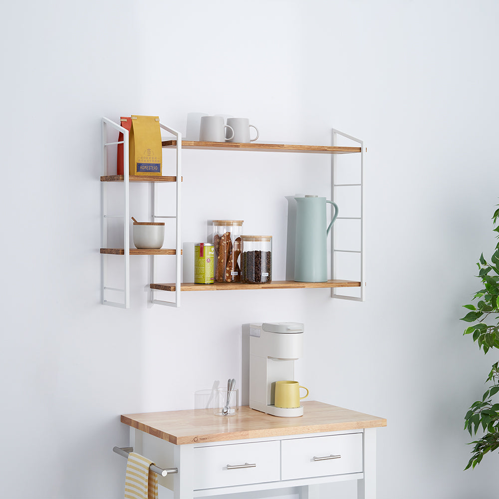 A two-tiered, wall-mounted shelf with a white metal frame and light wood shelves holds various coffee and tea supplies, including canisters, bags, mugs, and a thermos. Below the shelf, a white coffee maker sits on a wooden kitchen cart or island.